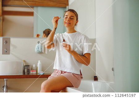 Pretty female applying serum on face moisturizing and caring for skin standing in bathroom 112645197