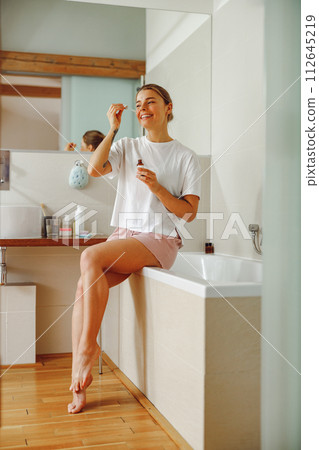 Attractive female applying serum on face moisturizing and caring for skin standing in bathroom Attractive female applying serum on face moisturizing and caring for skin standing in bathroom 112645219