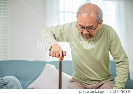 Elderly man suffering from knee pain ache holding handle of cane, senior old man typing to stand up from sofa alone with walking cane stick to walk at home, retirement medical healthcare concept Elderly man suffering from knee pain ache holding handle of cane, senior old man typing to stand up from sofa alone with walking cane stick to walk at home, retirement medical healthcare concept 112645689
