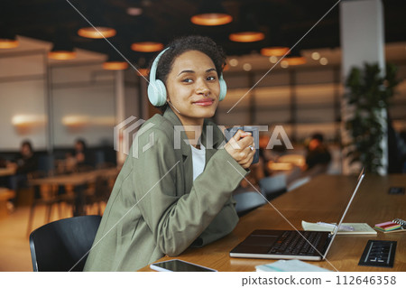 Woman with headphones sits at table with laptop, coffee, smiling in nice modern coworking office 112646358