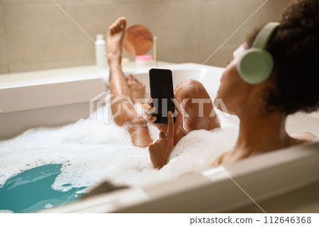 a woman is taking a bath in a bathtub while listening to music on her phone a woman is taking a bath in a bathtub while listening to music on her phone 112646368
