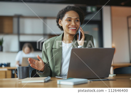 A woman sitting at a wood desk with a laptop, talking on her cell phone, smiling in nice modern coworking office 112646400