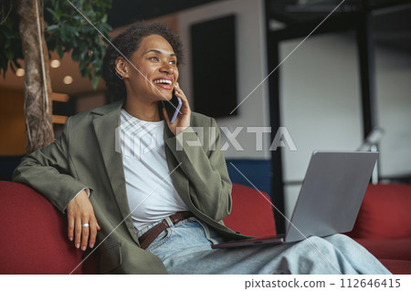 Woman in formal wear sitting on couch, talking on cell phone, using laptop in nice modern coworking office 112646415