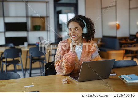 A woman with headphones smiles at a laptop on a wood table 112646510
