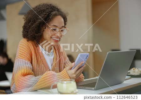 A woman is smiling at her laptop and cell phone on the table 112646523