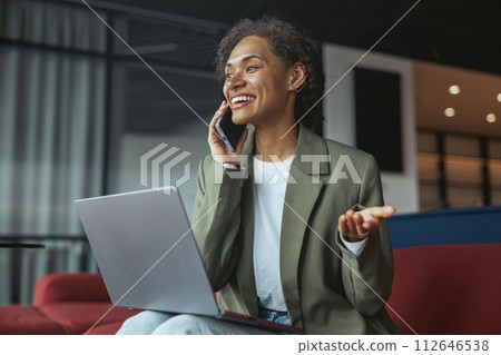 a woman is sitting on a couch talking on a cell phone while using a laptop computer in nice modern coworking office a woman is sitting on a couch talking on a cell phone while using a laptop computer in nice modern coworking office 112646538