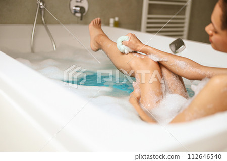 A woman is leisurely bathing, lathering soap on her arms and legs in a bathtub A woman is leisurely bathing, lathering soap on her arms and legs in a bathtub 112646540