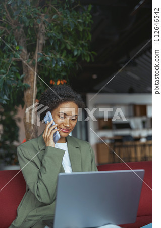 Woman sitting in room using laptop and talking on cell phone in nice modern coworking office Woman sitting in room using laptop and talking on cell phone in nice modern coworking office 112646542
