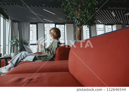 Woman sitting on red couch in nice modern coworking office, using laptop computer Woman sitting on red couch in nice modern coworking office, using laptop computer 112646544