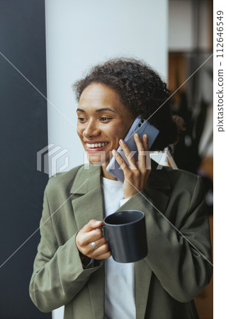 A woman smiles while holding a cup of coffee and talking on a mobile phone 112646549