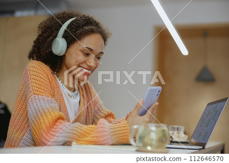 A young freelancer woman with headphones sitting happily at a table using laptop and cell phone A young freelancer woman with headphones sitting happily at a table using laptop and cell phone 112646570