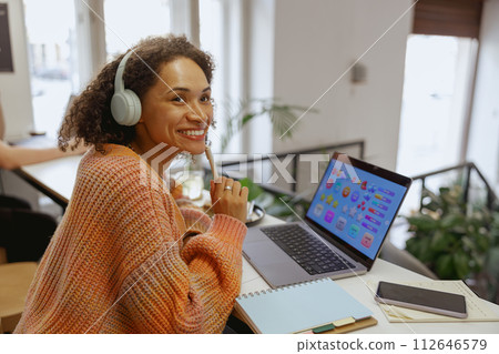 A woman with headphones sits at a table with a laptop, smiling 112646579