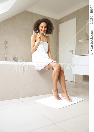 A woman in a towel sitting on the edge of the bathtub brushing her teeth 112646644