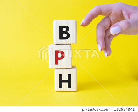 BPH - Benign Prostatic Hyperplasia symbol. Wooden cubes with word BPH. Businessman hand. Beautiful yellow background. Medical and BPH concept. Copy space. 112654841