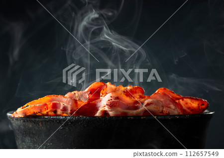 Fried steaming bacon slices in an old black pan. Fried steaming bacon slices in an old black pan. 112657549