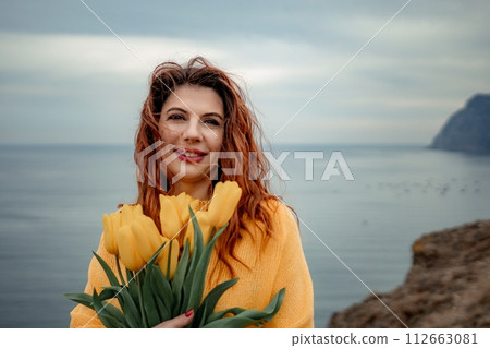 Portrait of a happy woman with hair flying in the wind against the backdrop of mountains and sea. Holding a bouquet of yellow tulips in her hands, wearing a yellow sweater 112663081