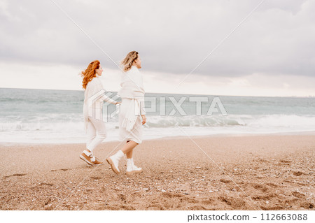 Women sea walk friendship spring. Two girlfriends, redhead and blonde, middle-aged walk along the sandy beach of the sea, dressed in white clothes. Against the backdrop of a cloudy sky and the winter 112663088