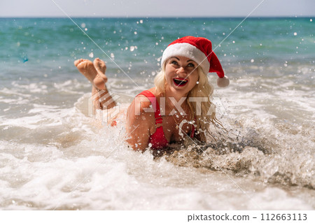 Female beach Santa hat wave coast. beach relaxation seaside. A woman in a red swimsuit enjoying her time on the beach, lying on the sand and being covered by a wave. Female beach Santa hat wave coast. beach relaxation seaside. A woman in a red swimsuit enjoying her time on the beach, lying on the sand and being covered by a wave. 112663113