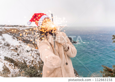 Outdoor winter portrait of happy smiling woman, light faux fur coat holding heart sparkler, posing against sea and snow background Outdoor winter portrait of happy smiling woman, light faux fur coat holding heart sparkler, posing against sea and snow background 112663158