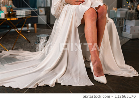Pregnant Women legs christmas tree. Cropped photo of beautiful elegant female legs in white high-heeled shoes and a white dress, a woman sits against the background of a Christmas tree with gifts. 112663170