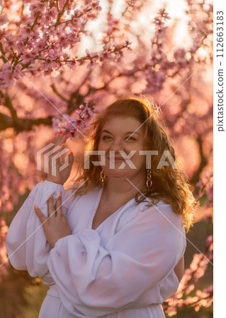 Woman blooming peach orchard. Against the backdrop of a picturesque peach orchard, a woman in a long white dress enjoys a peaceful walk in the park, surrounded by the beauty of nature. Woman blooming peach orchard. Against the backdrop of a picturesque peach orchard, a woman in a long white dress enjoys a peaceful walk in the park, surrounded by the beauty of nature. 112663183
