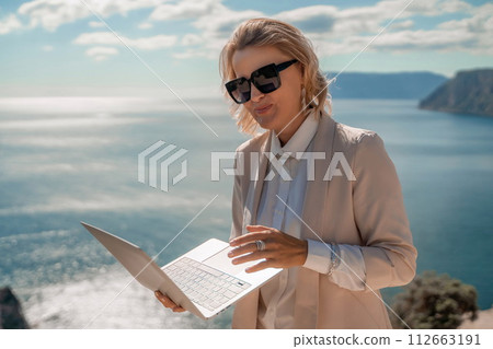 Freelance women sea. She is working on the computer. Good looking middle aged woman typing on a laptop keyboard outdoors with a beautiful sea view. The concept of remote work. Freelance women sea. She is working on the computer. Good looking middle aged woman typing on a laptop keyboard outdoors with a beautiful sea view. The concept of remote work. 112663191