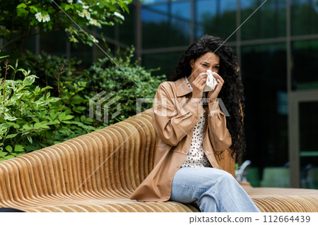 A woman sits on an outdoor bench, blowing her nose with a tissue, possibly suffering from cold or allergies, in a natural, urban setting. 112664439