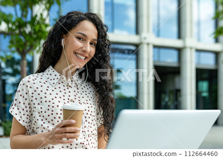 Smiling young woman with headphones and coffee taking a break from work on her laptop outside a modern office building. 112664460