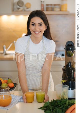 Beautiful woman in the kitchen finish make a fresh juice. Two glasses of different beverage on the table Beautiful woman in the kitchen finish make a fresh juice. Two glasses of different beverage on the table 112665207
