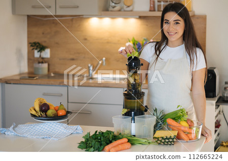 Young woman put celery into juicer. Process of making healthy drink. Place for text. Female look into camera Young woman put celery into juicer. Process of making healthy drink. Place for text. Female look into camera 112665224