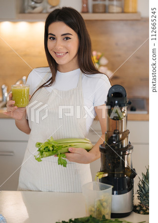 Portrait of beautiful and smiled female in apron standing in the kitchen. Woman hold glass with green healthy beverage in one hand and celery in another. Detox diet. Homemade juice. Vegetarian Portrait of beautiful and smiled female in apron standing in the kitchen. Woman hold glass with green healthy beverage in one hand and celery in another. Detox diet. Homemade juice. Vegetarian 112665245