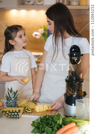 Little girl helps her mom in the kitchen. Women slice ananas for fresh homemade juice Little girl helps her mom in the kitchen. Women slice ananas for fresh homemade juice 112665252