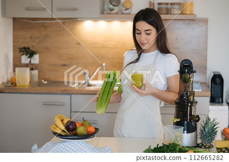 Portrait of beautiful and young female in apron standing in the kitchen. Woman hold glass with green healthy beverage in one hand and celery in another. Detox diet. Homemade juice. Vegetarian 112665282