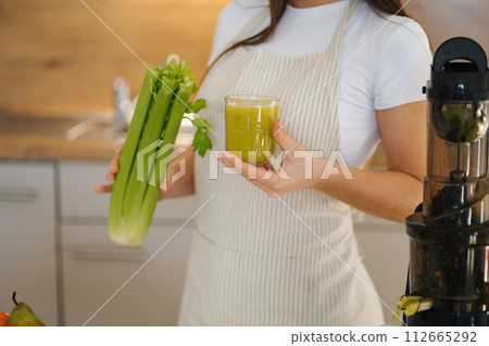 Close-up young female in apron standing in the kitchen. Woman hold glass with green healthy beverage in one hand and celery in another. Detox diet. Homemade juice. Vegetarian. Juicer. 112665292