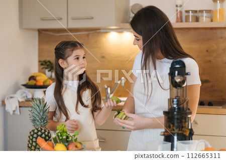 Adorable little girl helping her mom to make fresh juice at home. Two females in apron. Fruits and vegetables. Homemade vegan beverage Adorable little girl helping her mom to make fresh juice at home. Two females in apron. Fruits and vegetables. Homemade vegan beverage 112665311
