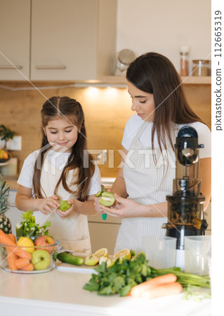Adorable little girl helping her mom to make fresh juice at home. Two females in apron. Fruits and vegetables. Homemade vegan beverage  112665319