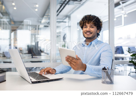Confident young businessman with curly hair smiling while using a tablet and laptop at his white desk in a bright contemporary office space. Confident young businessman with curly hair smiling while using a tablet and laptop at his white desk in a bright contemporary office space. 112667479