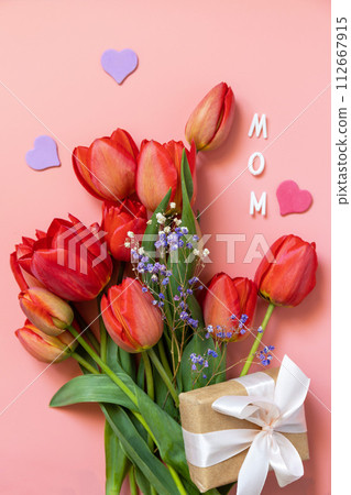 Red tulips with gift box and small hearts, "mom" handwritten text on pastel pink background with empty space. 112667915