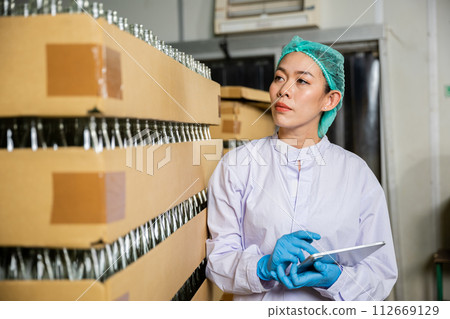 Within a beverage factory a QC woman checks products on the conveyor belt using a digital tablet. Her role involves quality control while examining the bottling line for liquid manufacturing. Within a beverage factory a QC woman checks products on the conveyor belt using a digital tablet. Her role involves quality control while examining the bottling line for liquid manufacturing. 112669129