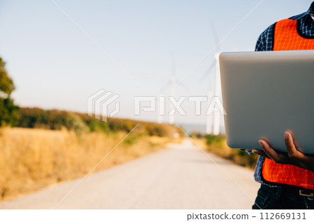 Engineer laptop in hand assesses wind turbines. Service technician ensures efficiency. Focused on innovation quality and landscape development in windmill service. Engineer laptop in hand assesses wind turbines. Service technician ensures efficiency. Focused on innovation quality and landscape development in windmill service. 112669131