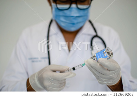 Doctors are experimenting with vaccination against the coronavirus. Scientists are using a syringe. A man wearing a mask holds a syringe and a vaccine bottle. Concept of the laboratory vaccine trial. Doctors are experimenting with vaccination against the coronavirus. Scientists are using a syringe. A man wearing a mask holds a syringe and a vaccine bottle. Concept of the laboratory vaccine trial. 112669202