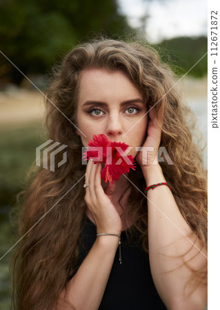 Elegant woman poses with red blossom, nature backdrop. Female holds flower in mouth, symbolize beauty, femininity. In portrait, captivating gaze, serene atmosphere, outdoor setting, flower in lip. Elegant woman poses with red blossom, nature backdrop. Female holds flower in mouth, symbolize beauty, femininity. In portrait, captivating gaze, serene atmosphere, outdoor setting, flower in lip. 112671872