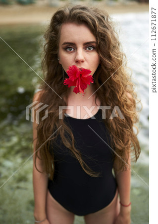Attractive woman in swimsuit holds red hibiscus in mouth by water. Natural beauty, wellness, summer fashion concept. Feminine allure with tropical flora. Lady enjoys beach lifestyle, relaxation. Attractive woman in swimsuit holds red hibiscus in mouth by water. Natural beauty, wellness, summer fashion concept. Feminine allure with tropical flora. Lady enjoys beach lifestyle, relaxation. 112671877