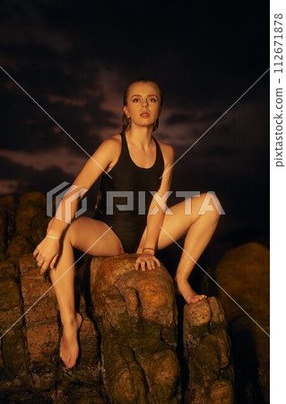 Woman in black swimsuit poses on rocky terrain at sunset. Model showcases sleek swimwear design, fashion shoot by ocean. Twilight lighting enhances elegant beachwear attire, serene summer evening vibe Woman in black swimsuit poses on rocky terrain at sunset. Model showcases sleek swimwear design, fashion shoot by ocean. Twilight lighting enhances elegant beachwear attire, serene summer evening vibe 112671878