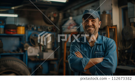 Confident Caucasian male auto mechanic in blue uniform stands with arms crossed in auto repair shop. 112673382