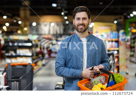 An adult male holds a shopping basket with fresh produce as he shops in the grocery section of a supermarket. 112673724