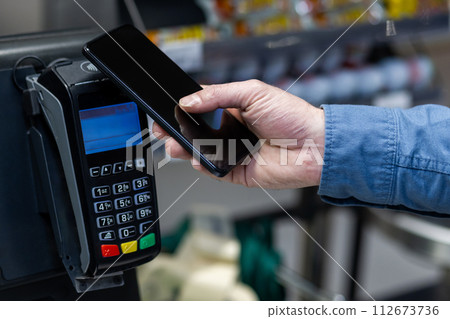 Close-up of a male hand holding a smartphone to a card reader for mobile payment in a retail store setting. 112673736