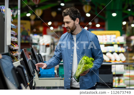 Adult male in a blue shirt using a self-checkout machine in a supermarket, holding fresh lettuce. 112673749