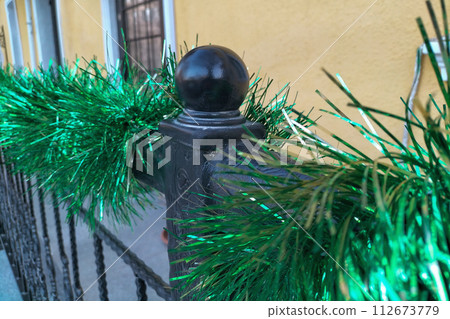 A black metal fence with decorative ornaments and lines is decorated with green silver festive tinsel. Creating a festive atmosphere on the streets of the city A black metal fence with decorative ornaments and lines is decorated with green silver festive tinsel. Creating a festive atmosphere on the streets of the city 112673779