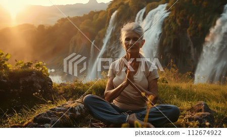 beautiful elderly woman doing yoga in nature against the background of waterfall and sunset. 112674922
